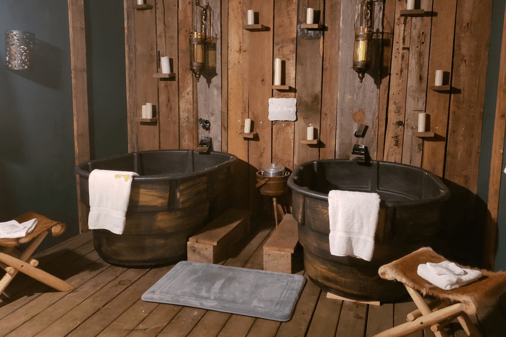 A rustic bathroom featuring two metal bathtubs, wooden walls, and candle displays.