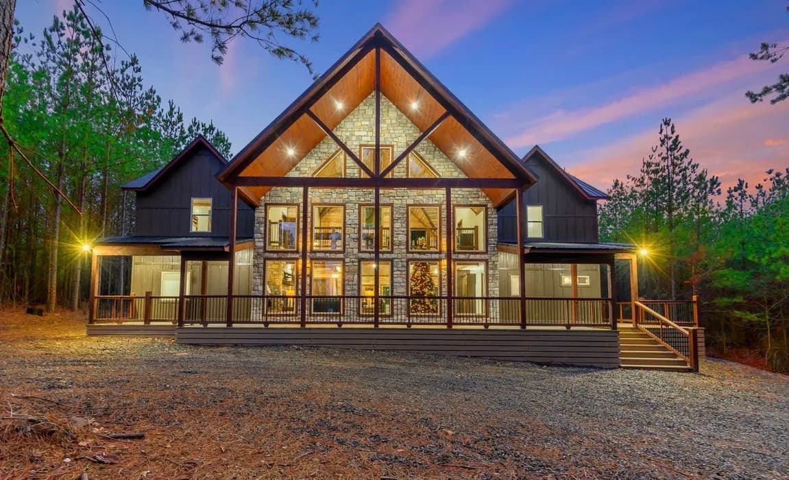 A modern chalet-style house with large windows, surrounded by pine trees at sunset.