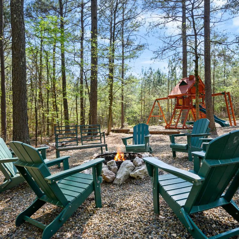 A cozy outdoor seating area with green chairs surrounding a fire pit, set among tall trees and a playground in the background.