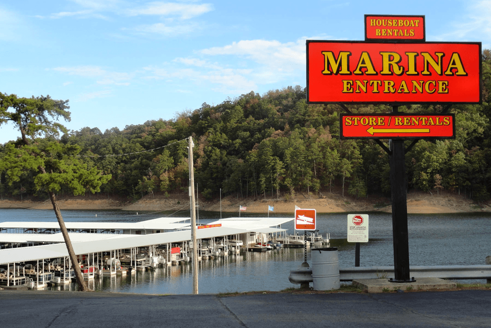 A bright red sign for a marina entrance, indicating houseboat rentals and a store, near a calm lake and wooded hillside.
